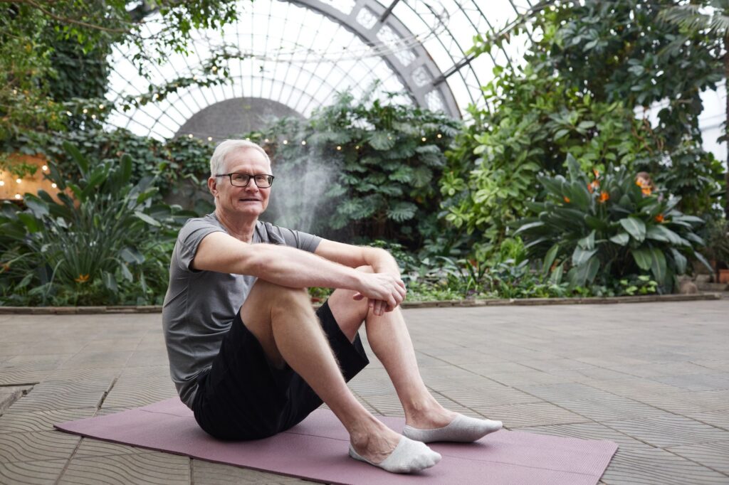 Older man enjoying yoga session on a mat in a lush botanical garden.