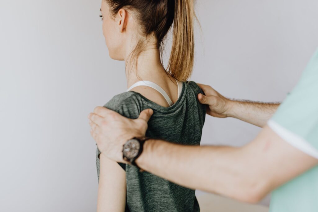 Back view of crop faceless orthopedist in uniform and wristwatch examining shoulder joints of slim female patient in clinic